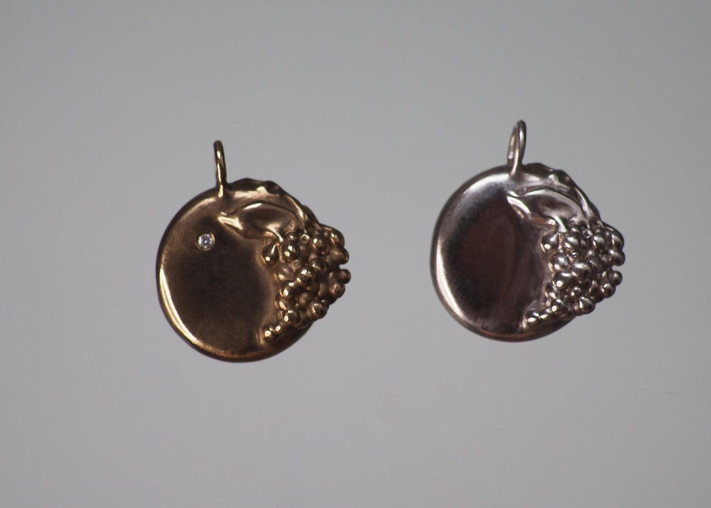 Two grape-detailed medallion pendants in solid gold and sterling silver on a dark gray background