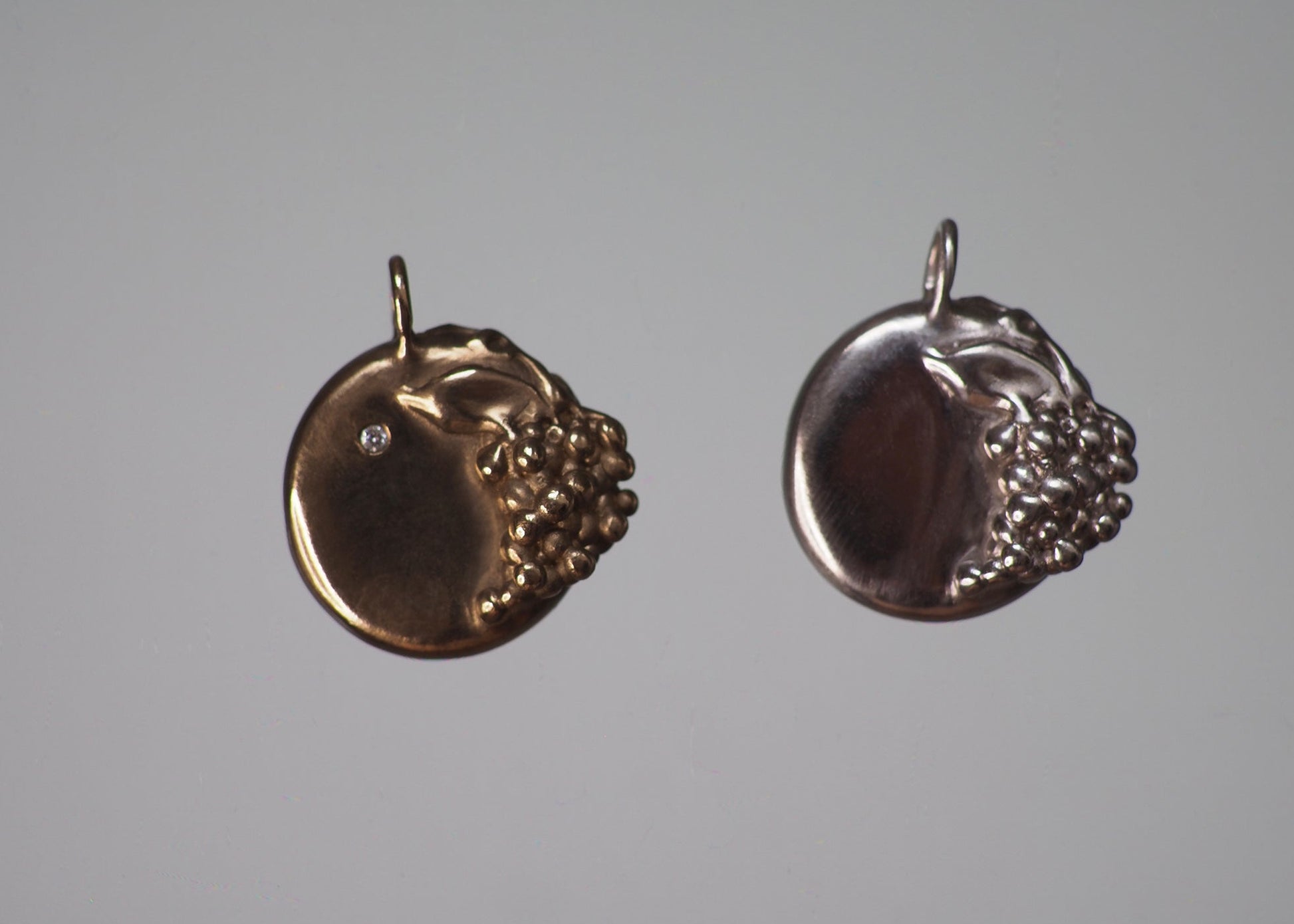 Two grape-detailed medallion pendants in solid gold and sterling silver on a dark gray background