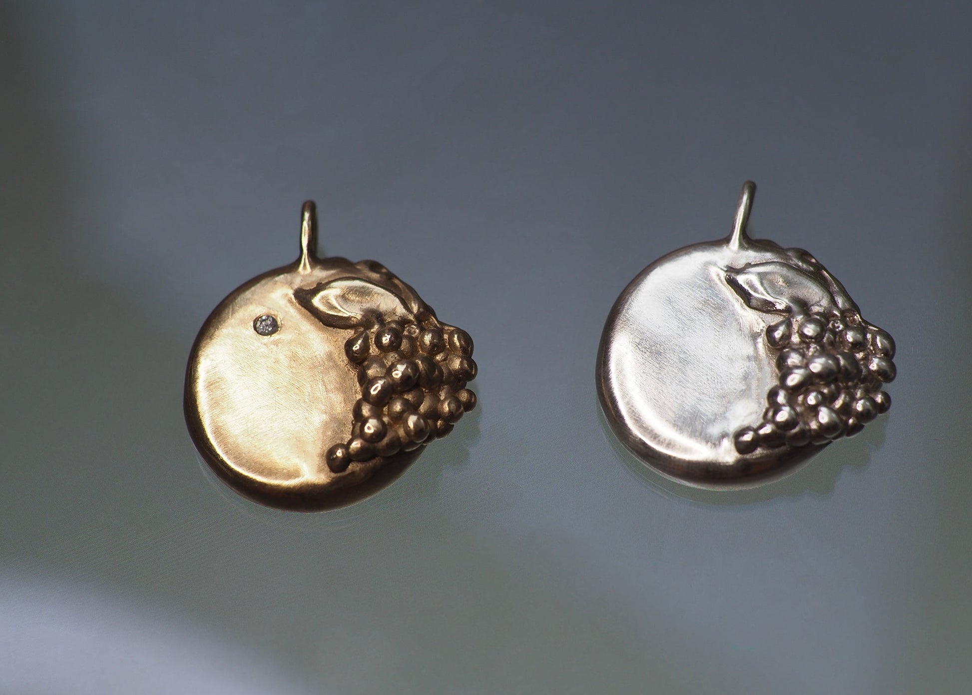 Two grape-detailed medallion pendants in solid gold and sterling silver on a dark gray background